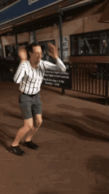 a man in a plaid shirt and shorts is dancing on a sidewalk in front of a sign that says " bowling open "