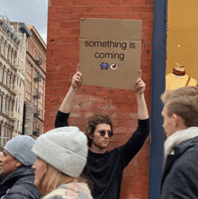 a man holds up a cardboard sign that says something is coming