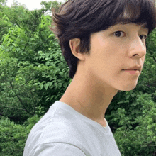 a young man with short hair is wearing a white shirt and standing in front of a forest .