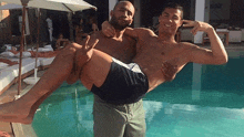 a shirtless man is carrying another shirtless man in his arms by a swimming pool .