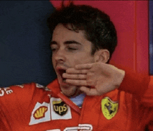 a man in a red ferrari jacket is yawning with his hand on his mouth .