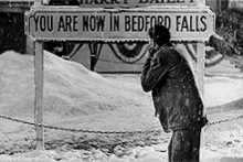 a black and white photo of a man standing in front of a sign that says you are now in bedford falls