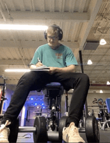 a man wearing headphones sits on a bench in a gym writing in a notebook