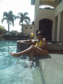 a woman in a yellow bikini sits on the edge of a swimming pool holding a drink