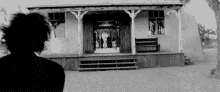 a black and white photo of a man standing in front of a house with people standing on the porch .