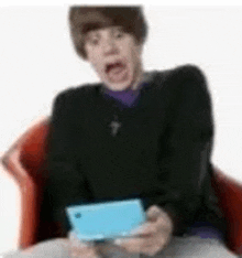 a man is sitting in a chair holding a blue nintendo 3ds and making a funny face .