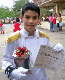 a boy in a white uniform holds a diploma and a gift