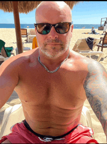 a shirtless man wearing sunglasses and a chain sits on a beach