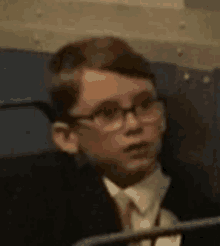 a young boy wearing glasses and a suit is sitting in a dark room .