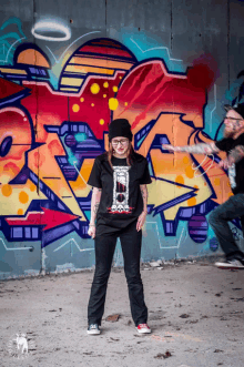 a woman in a black shirt stands in front of a wall with graffiti on it that says ' x '