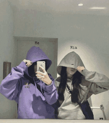 a girl in a purple hoodie is taking a picture of herself with another girl in a grey hoodie