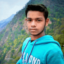 a young man wearing a blue lloyd hoodie stands in front of a mountain