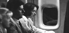 a black and white photo of a man and a woman sitting on an airplane looking out the window .