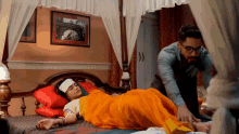 a man standing next to a woman laying on a bed with an orange blanket