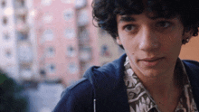 a young man with curly hair looks at the camera with a pink building in the background