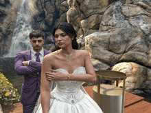 a man in a purple suit stands next to a woman in a white dress in front of a waterfall