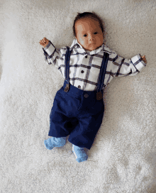 a baby wearing a plaid shirt and suspenders is laying on a white blanket