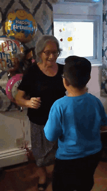 a boy in a blue shirt is dancing with an older woman in a black shirt with a happy birthday balloon in the background