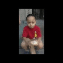 a young boy in a red shirt is sitting on the ground holding a bottle .