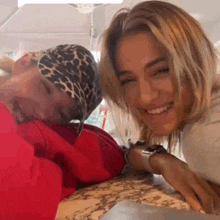 two women are sitting at a table and smiling at the camera . one of the women is wearing a leopard print bandana .