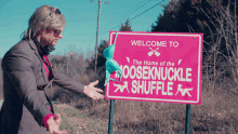 a man in a suit stands in front of a pink sign that says welcome to the home of the nooseknuckle shuffle