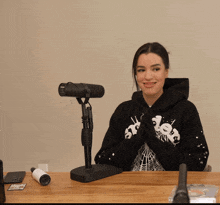 a woman wearing a black hoodie with a spider web on it sits at a desk in front of a microphone