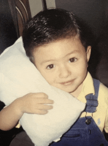 a young boy in blue overalls is holding a white pillow and smiling