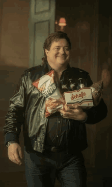 a man in a leather jacket is holding a bag of potato chips and a six pack of schafe beer