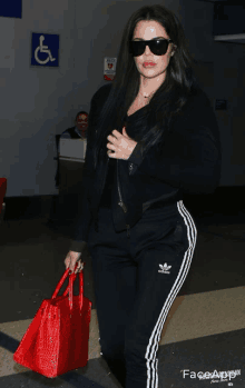 a woman wearing sunglasses and black adidas sweatpants holds a red bag