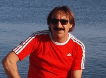 a man wearing sunglasses and a red adidas shirt stands in front of the water