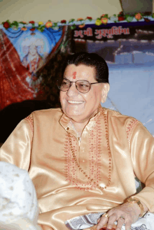 a man wearing glasses and a gold shirt smiles in front of a sign that says ' aarti '