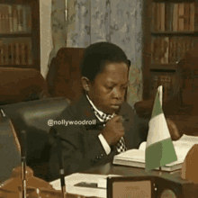 a man in a suit and tie sits at a desk with a small nigerian flag in front of him