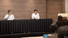 two men are sitting at a table talking into microphones while a group of people sit in front of them .