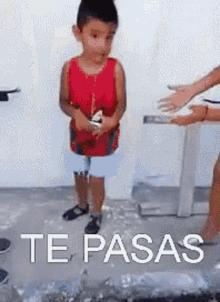 a little boy in a red tank top is standing in front of a table with the words te pasas written on it .