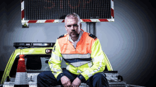 a man in a high visibility jacket sits on the back of a truck in front of a sign that says stop