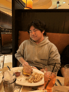 a man wearing a grey essentials hoodie sits at a table with a plate of food