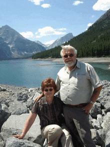 a man and woman pose for a picture in front of a lake with mountains in the background