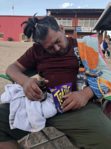 a man sits in a chair with a bag of takis and a bottle of beer