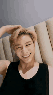 a young man in a black tank top smiles while sitting on a bed