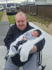 a man in a wheelchair holds a baby in his arms with a car parked in the background