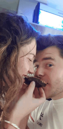 a woman is feeding a man a piece of chocolate cake while wearing a shirt that says ' xkri ' on it