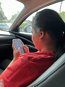 a woman in a red shirt is sitting in a car looking at her cell phone