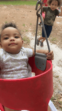 a baby is sitting in a red swing while another child stands behind him