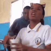 a girl in a school uniform is sitting at a desk in a classroom with a cup in her hand .