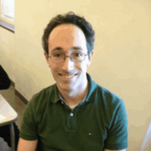 a man wearing glasses and a green shirt is smiling for the camera while sitting at a table .