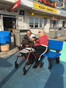 a man and woman sit on a blue bench in front of a pizza cannoli store