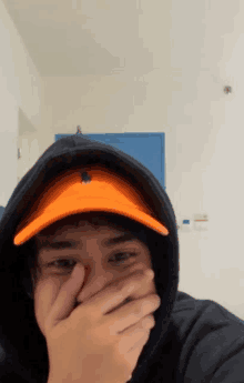 a man wearing an orange hat and a black hoodie covering his face with his hand