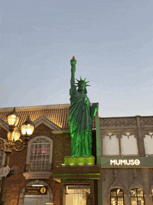 a green statue of the statue of liberty stands in front of a building that says mumuso