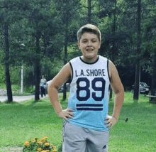 a young boy wearing a l.a. shore 89 jersey stands in a park with his hands on his hips .