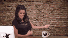 a woman is sitting at a desk in front of a brick wall with a laptop and a mug on it .